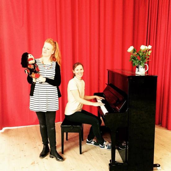 Et billede med to kvinder, hvor den ene spiller på klaver og den anden står med en dukke i hånden.