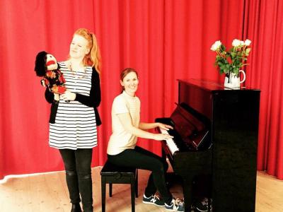 Et billede med to kvinder, hvor den ene spiller på klaver og den anden står med en dukke i hånden.