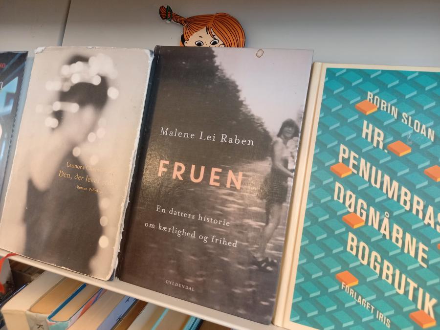 Foto af romanen 'Fruen' ved siden af nogle andre romaner på en skråhylde på Greve Bibliotek